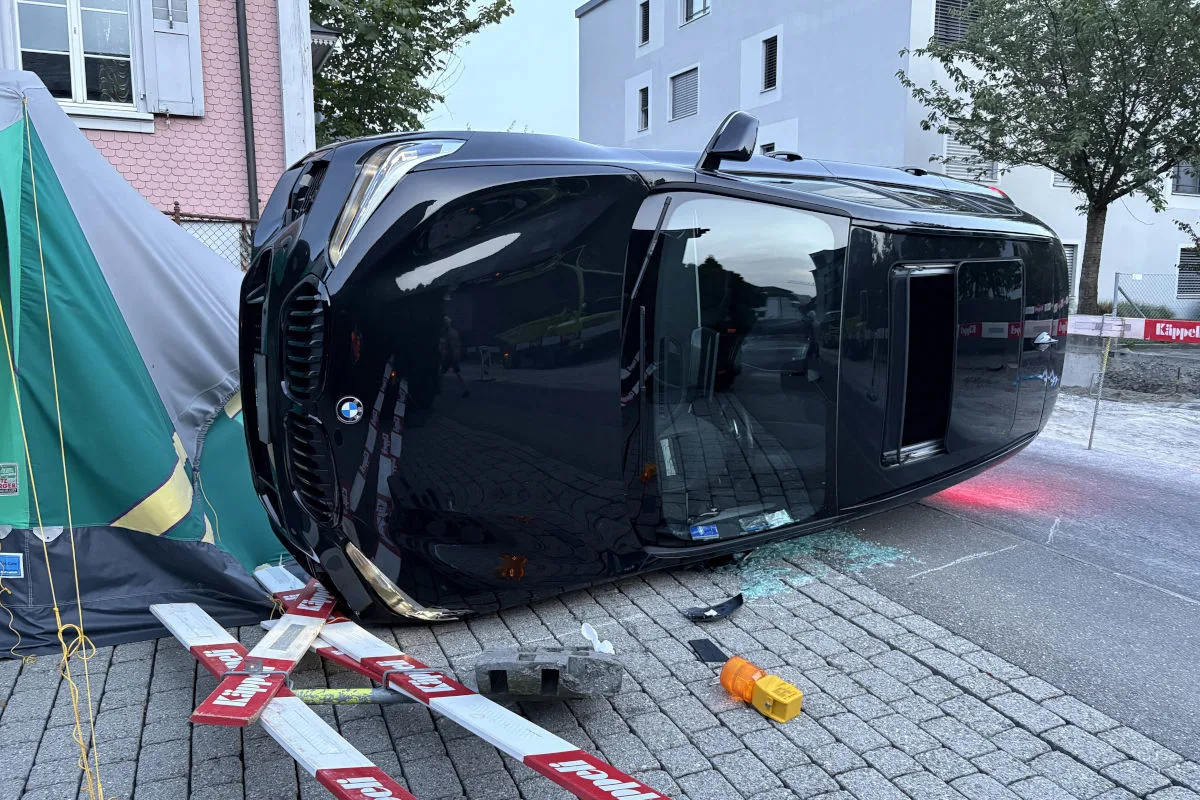 Ein schwarzer BMW ist auf die Seite gekippt und liegt auf Pflastersteinen vor einem Gebäude. Neben dem Auto sind Absperrungen und ein Zelt sichtbar.