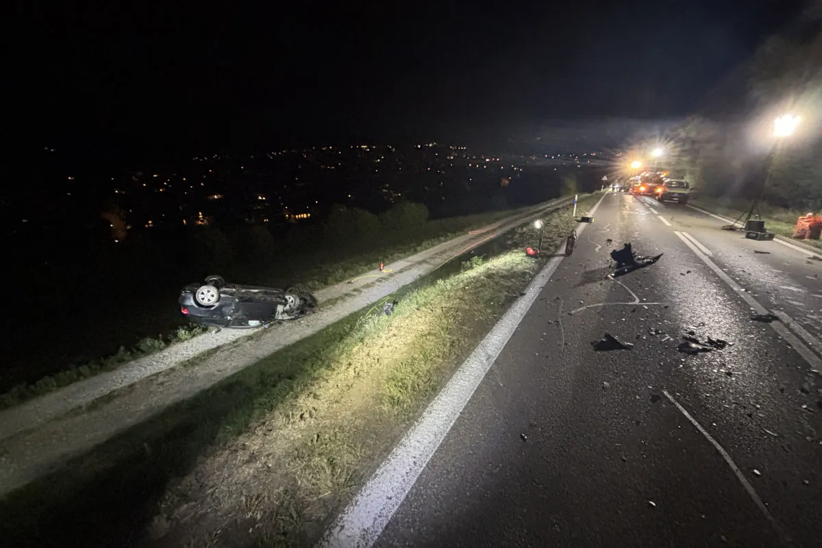 Ein Auto liegt auf dem Dach neben einer Straße, die bei Nacht beleuchtet ist. Trümmer sind auf der Fahrbahn sichtbar.