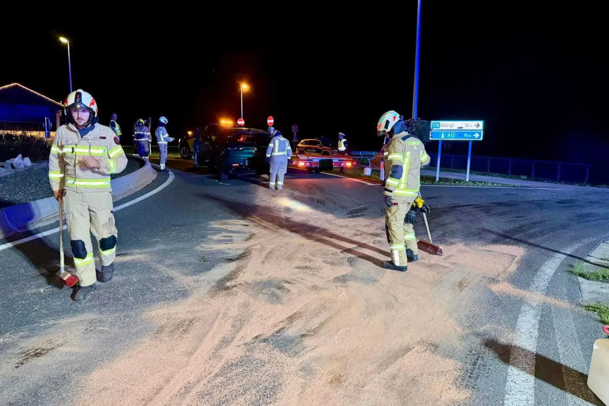 Mehrere Feuerwehrleute arbeiten nachts auf einer Straße, um ausgelaufene Flüssigkeiten zu beseitigen. Im Hintergrund sind beschädigte Fahrzeuge und Licht von Einsatzfahrzeugen sichtbar.