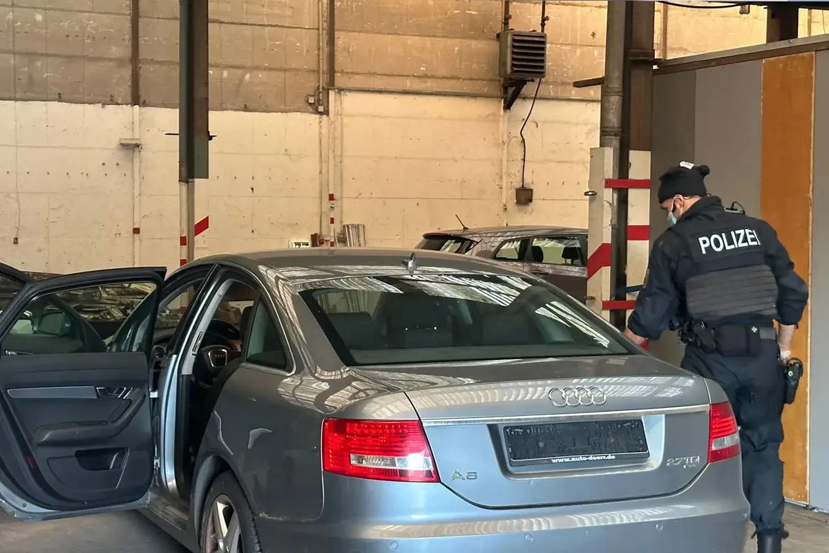 Ein Polizeiangehöriger inspiziert einen grauen Audi A6 in einer Fahrzeughalle. Die Umgebung wirkt industriell.