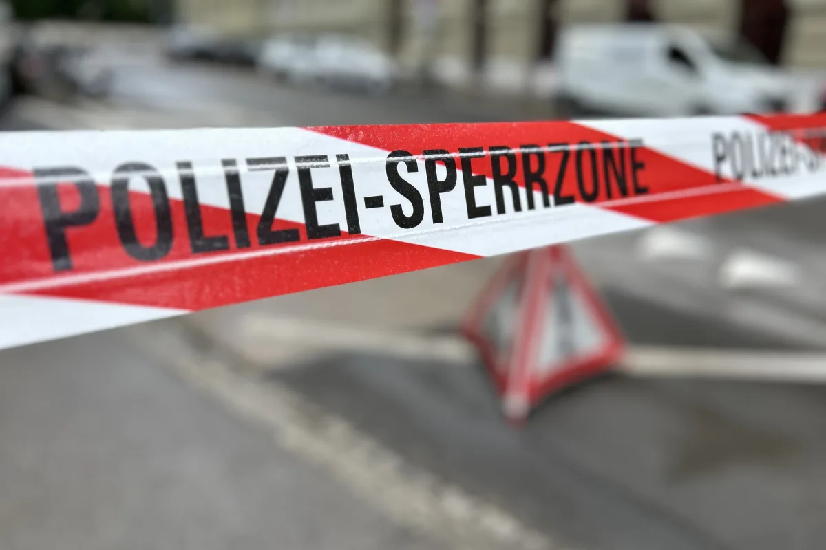 Ein Polizeiabsperrband in Rot-Weiß markiert eine Sperrzone auf einer Straße. Im Hintergrund sind unscharfe Gebäude und Fahrzeuge sichtbar.