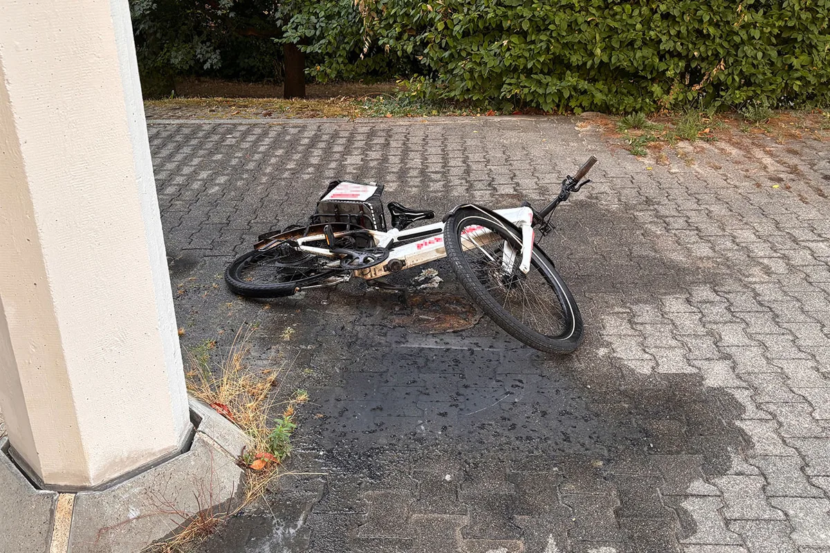 Ein umgestürztes E-Bike liegt auf einem gepflasterten Bereich neben einem weißen Pfeiler. Büsche sind im Hintergrund sichtbar.