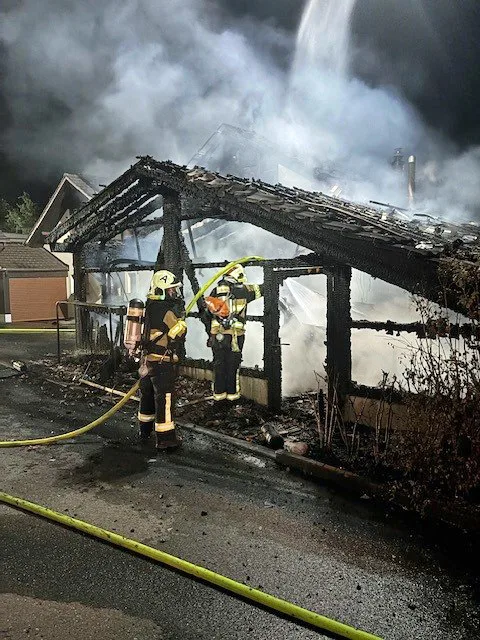 Zwei Feuerwehrleute in Schutzkleidung löschen ein Gebäude, das stark brennt. Dicker Rauch und Flammen sind sichtbar.