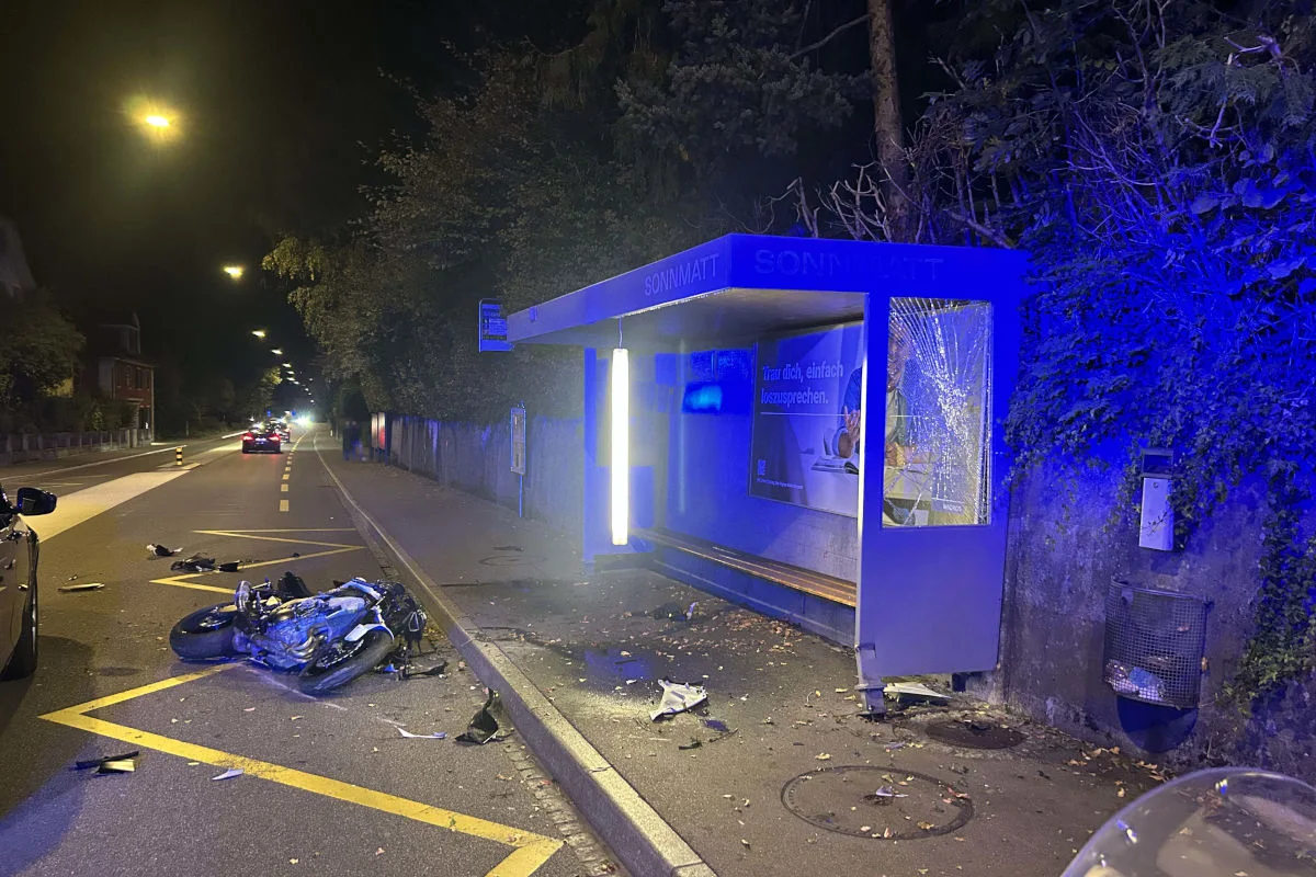 Ein beschädigtes Motorrad liegt vor einer Bushaltestelle, deren Scheibe zersplittert ist. Die Szene ist bei Nacht beleuchtet.