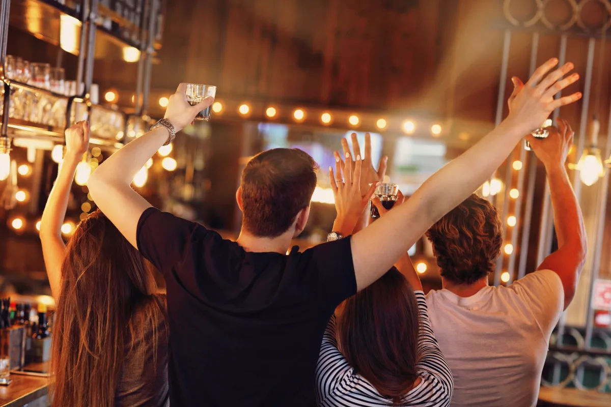 Eine Gruppe von Menschen hebt Gläser in einer Bar, umgeben von Lichtern und festlicher Atmosphäre.