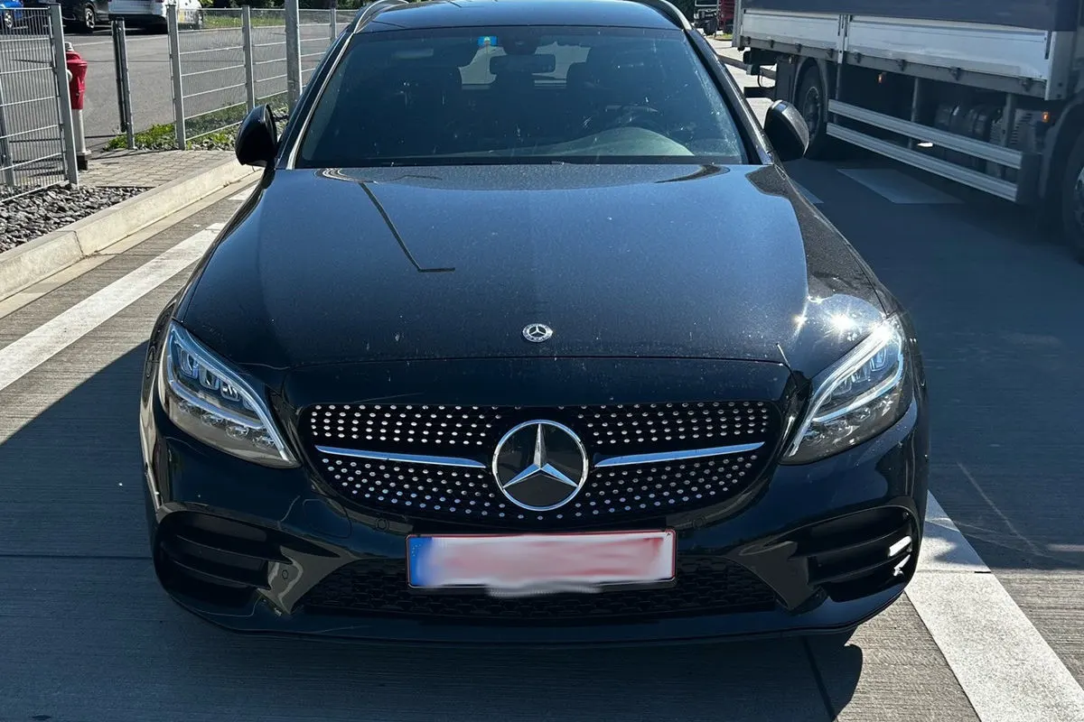 Der schwarze Mercedes steht frontal auf einem Rastplatz bei hellem Tageslicht. Ein LKW ist rechts im Hintergrund zu sehen.