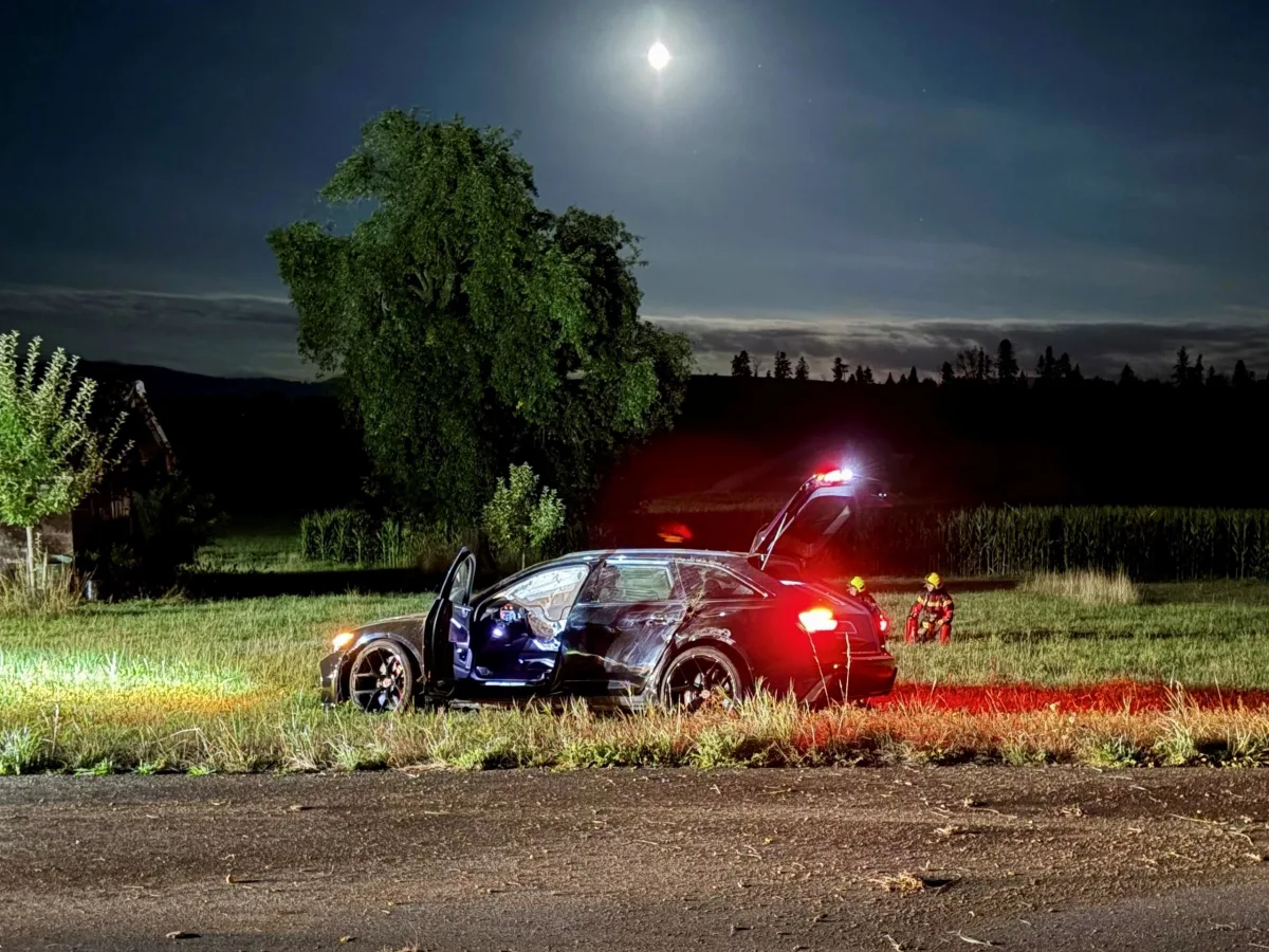 Ein Auto mit offener Tür und offenem Kofferraum steht bei Nacht auf einer Wiese, beleuchtet von Blaulicht. Zwei Einsatzkräfte sind in der Nähe zu sehen.