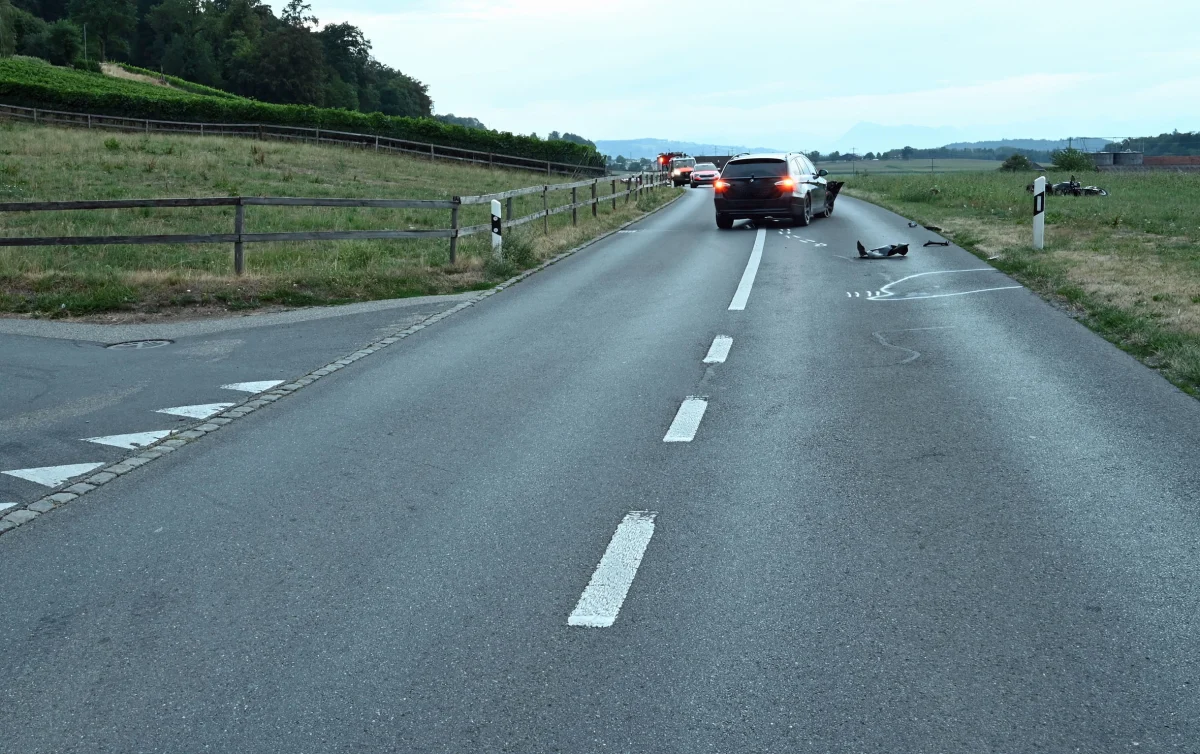 Ein Auto steht auf einer Landstraße, mit Motorradtrümmern auf der Fahrbahn und einer grasbewachsenen Umgebung.