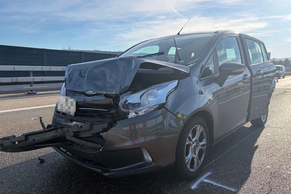 Ein Ford-Auto mit stark beschädigter Vorderseite steht auf einer Autobahn mit klarer Sicht und blauem Himmel.