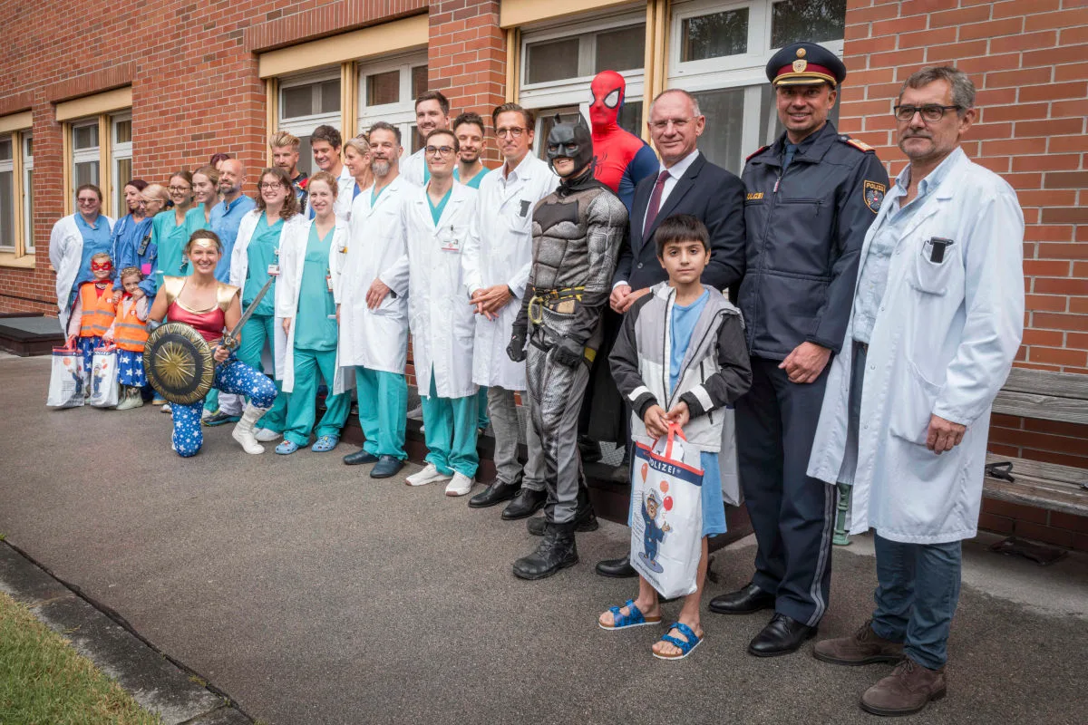 Eine Gruppe von erwachsenen Klinikmitarbeitern und ein Polizist stehen mit Kindern in Superheldenkostümen draußen vor einem Backsteingebäude. Einige tragen bunte Kostüme.