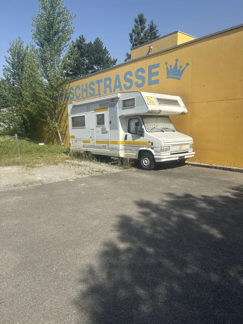 Ein weißes Wohnmobil steht auf einem Parkplatz vor einer gelben Wand mit dichterem Bewuchs und der blauen Aufschrift SCHSTRASSE.