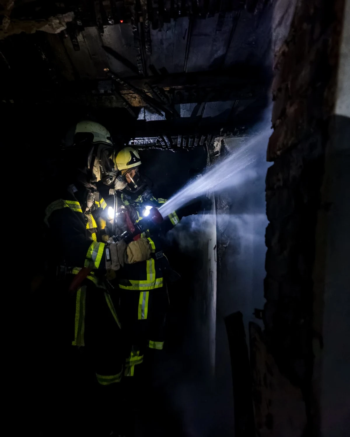 In einer dunklen Szenerie bekämpfen zwei Feuerwehrleute in vollständiger Schutzkleidung und mit Atemmasken einen Innenbrand, wobei sie einen starken Wasserstrahl auf die Brandstelle richten.