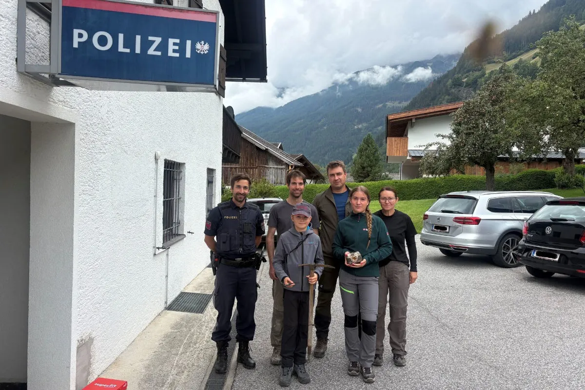 Eine Personengruppe steht vor einer Polizeistation. Im Hintergrund sind Berge und eine ländliche Gegend zu sehen.
