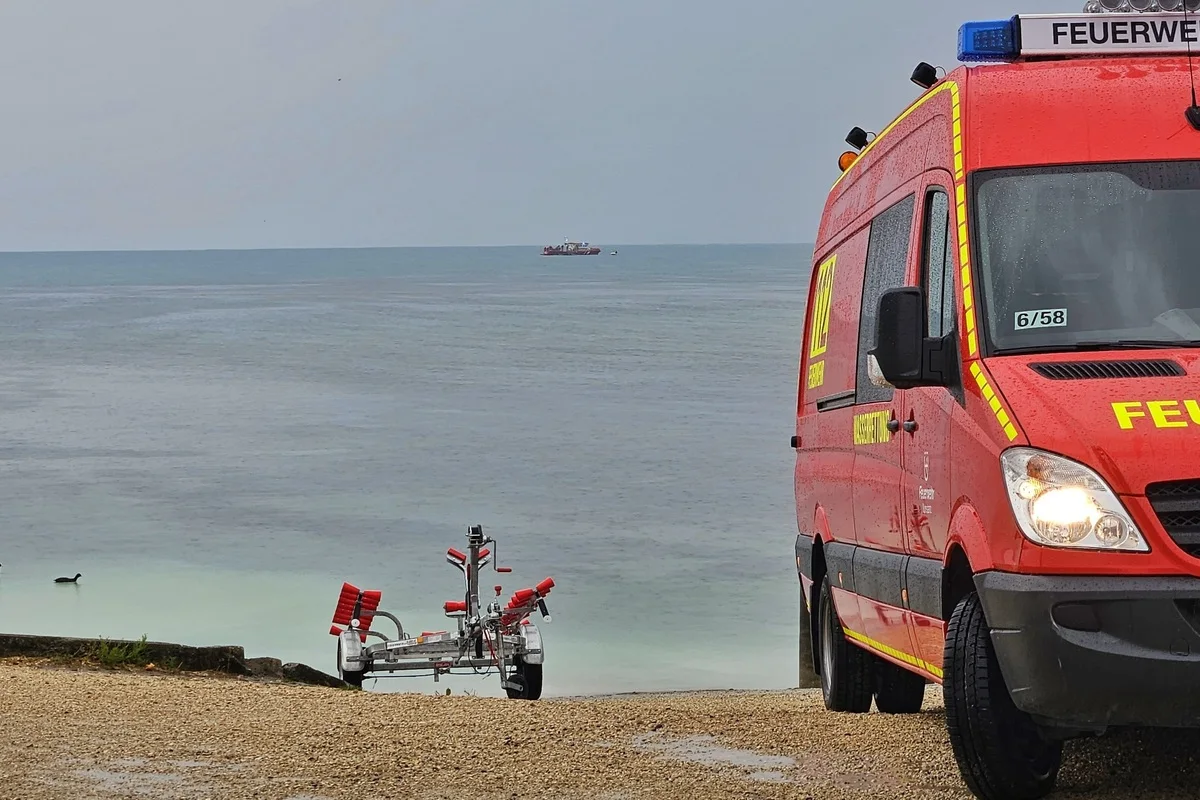 Ein rotes Feuerwehrfahrzeug steht auf kiesigem Boden nahe einem Strand. Ein leerer Bootsanhänger ist sichtbar, und im Hintergrund fährt ein Boot auf dem Wasser.