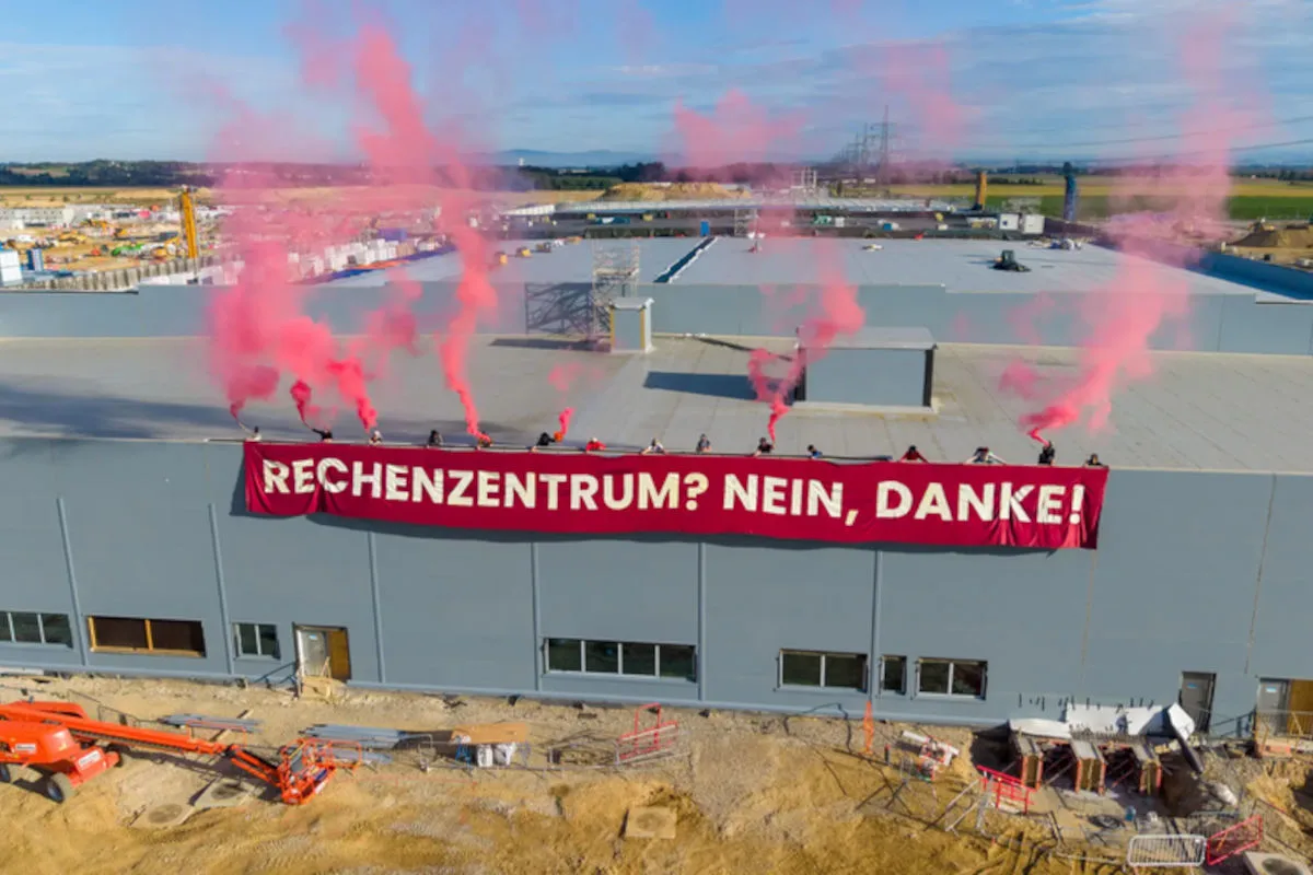 Eine Gruppe von Personen steht auf einem grauen Gebäudedach. Sie halten ein rotes Banner mit der Aufschrift 'Rechenzentrum? Nein, Danke!' Es steigt roter Rauch auf.