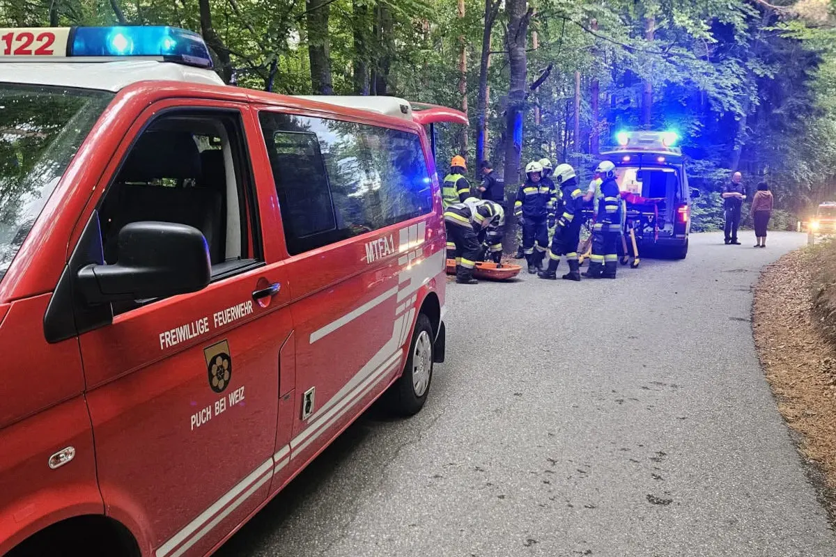 Ein rotes Feuerwehrfahrzeug der Freiwilligen Feuerwehr Puch bei Weiz und Einsatzkräfte auf einer Waldstraße. Mehrere Feuerwehrleute sind bei einer Rettungsaktion zu sehen.