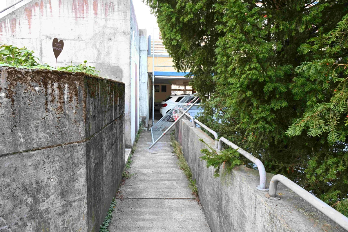 Ein Gehweg mit einem umgestürzten Metalltor vor einem geparkten weißen Auto. Rechts steht eine große grüne Hecke.