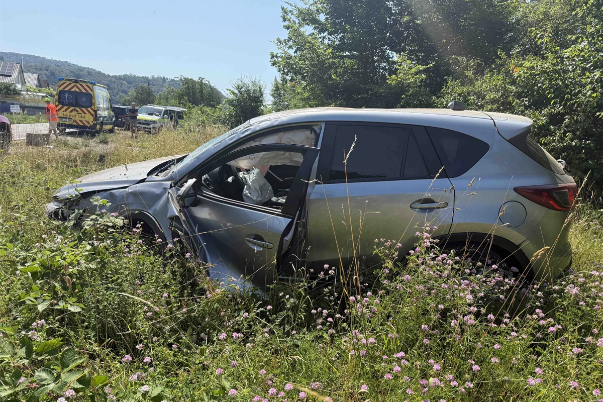 Ein silbernes Auto mit Beulen und offenem Airbag steht im hohen Gras neben einer Straße. Im Hintergrund sind Einsatzfahrzeuge sichtbar.