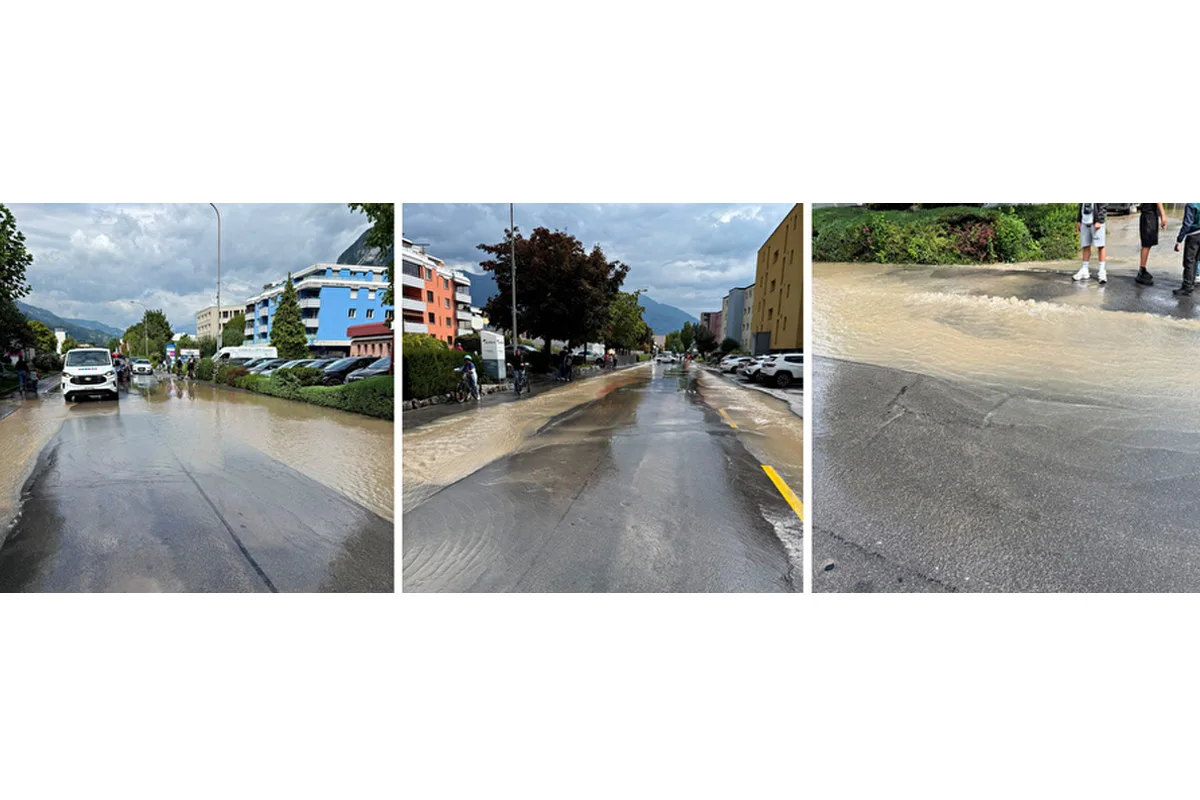 Eine überflutete Straße zeigt mehrere Autos, Fahrräder und angrenzende Gebäude. Der Himmel ist bewölkt und die Straße nass.