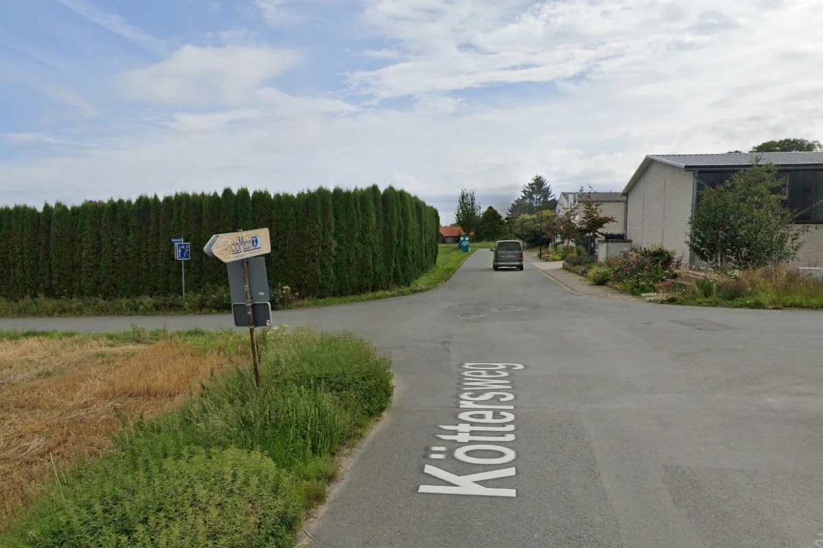 Ein silberner Van fährt auf dem Köttersweg entlang, gesäumt von Hecken und vereinzelten Häusern. Der Himmel ist bewölkt, die Straße wirkt ruhig und verlassen.