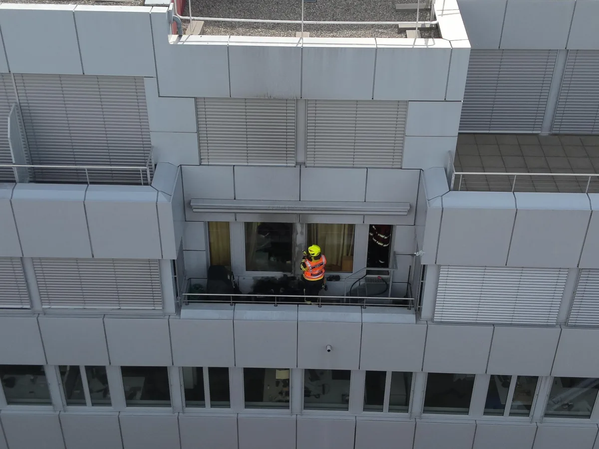 Ein Feuerwehrmann in Warnkleidung steht auf einem Balkon eines modernen Gebäudes mit metallischer Verkleidung. Im Hintergrund sind fast geschlossene Fensterläden sichtbar.