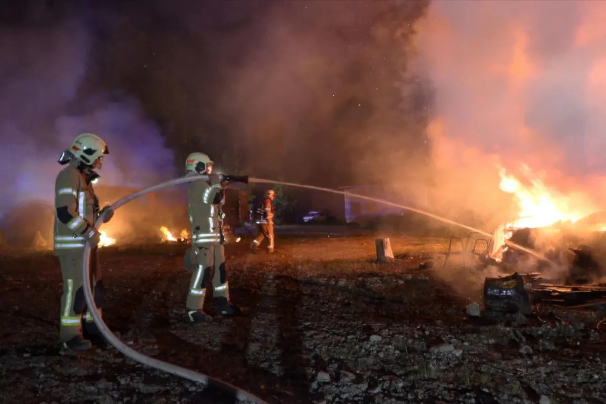 Drei Feuerwehrleute löschen bei Nacht mit Wasserschläuchen ein großes Feuer auf einer freien Fläche, dichte Rauchwolken steigen auf.