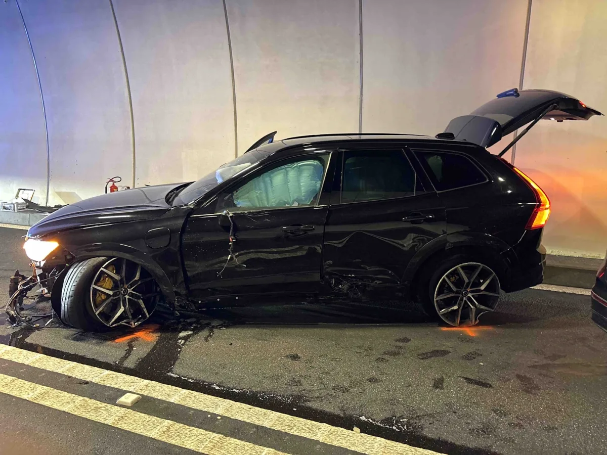 Ein schwarzes Auto mit erheblichen Schäden an der Vorderseite steht in einem Tunnel. Die Räder sind stark eingeschlagen, und es gibt Spuren von ausgetretenem Öl auf der Straße.