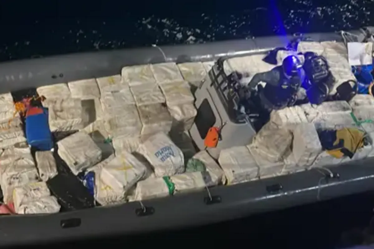 Ein großes Schlauchboot ist mit vielen rechteckigen Paketen beladen, auf einem Gewässer in der Dunkelheit. Eine Person in Uniform ist sichtbar.