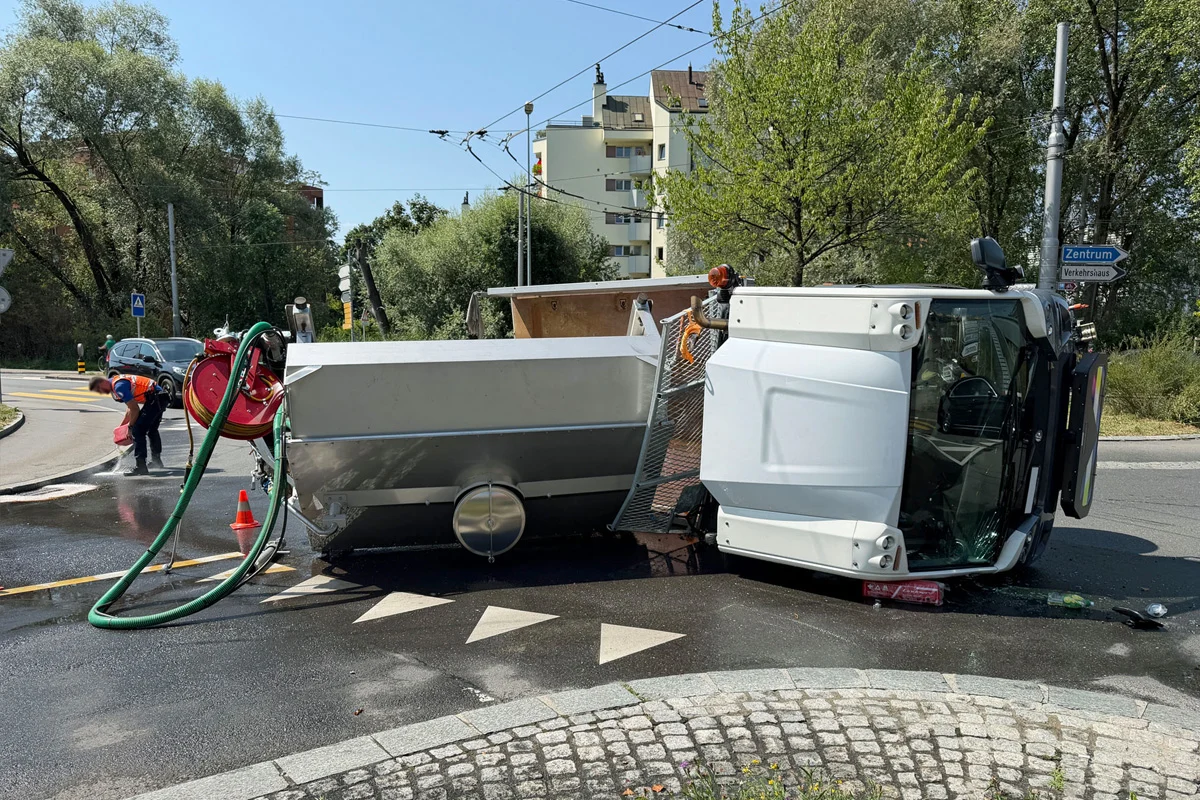 Ein weißer Lkw liegt auf der Seite und blockiert eine Kreuzung in einem Wohngebiet. Ein Arbeiter ist dabei, die verschmutzte Straße zu reinigen.