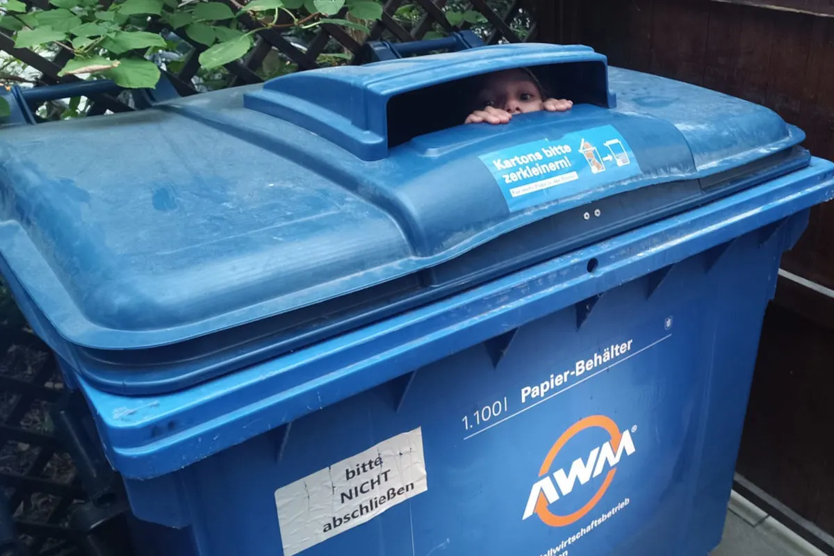 Ein kleines Kind schaut durch die Öffnung eines blauen Papiercontainers mit dem Logo 'AWM' und einem Aufkleber bei Tageslicht.