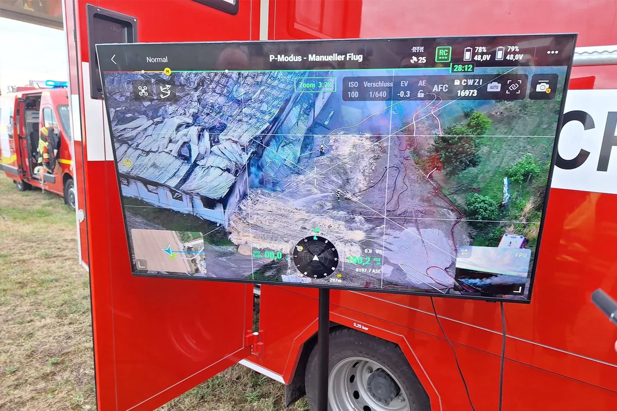Ein Monitor zeigt eine Luftaufnahme eines beschädigten Gebäudes, aufgenommen von einer Drohne. Daneben ist ein Einsatzfahrzeug der Feuerwehr sichtbar.
