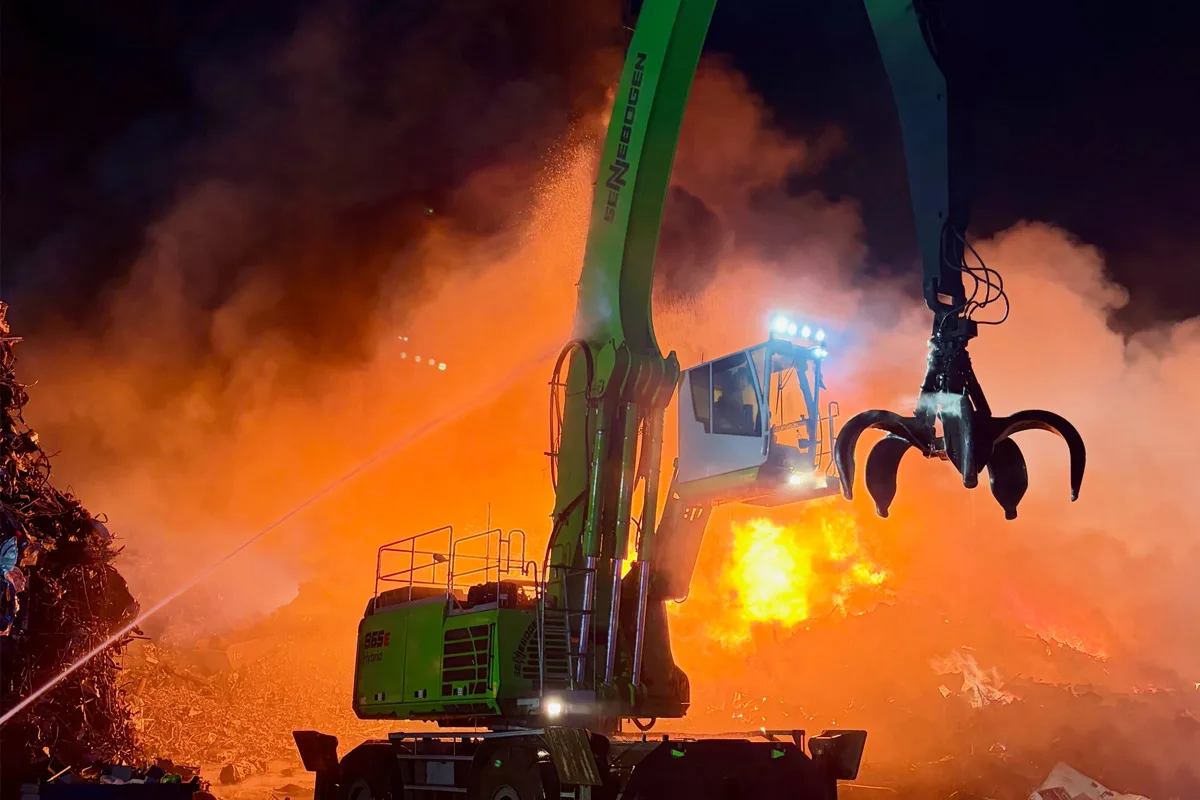 Ein großer, grüner Kran steht nachts auf einem Schrottplatz, greift in Flammen. Feuer und Rauch im Hintergrund.