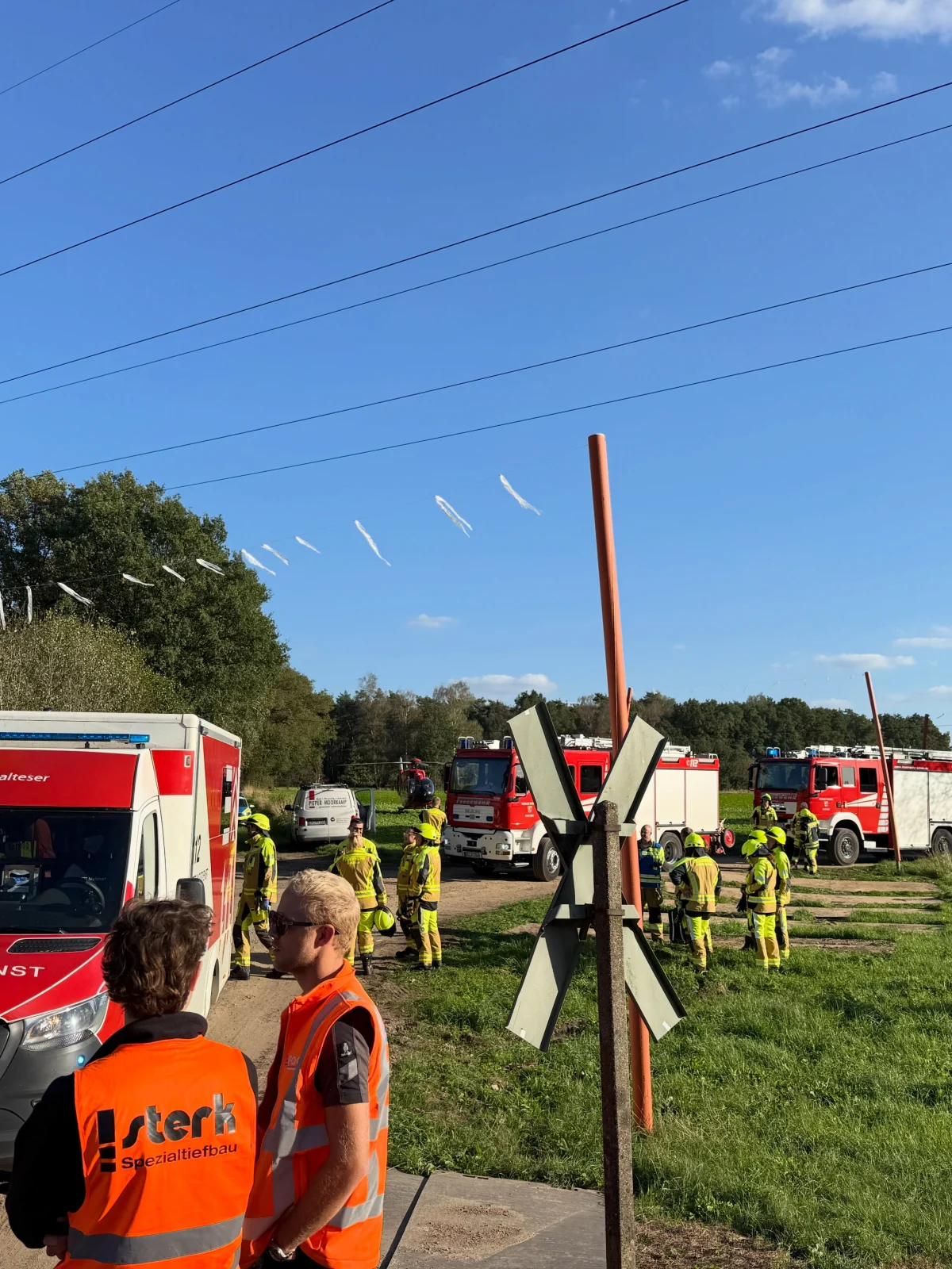 Mehrere Feuerwehrleute stehen in gelber Schutzkleidung neben Einsatzfahrzeugen auf einem ländlichen Weg bei sonnigem Wetter.