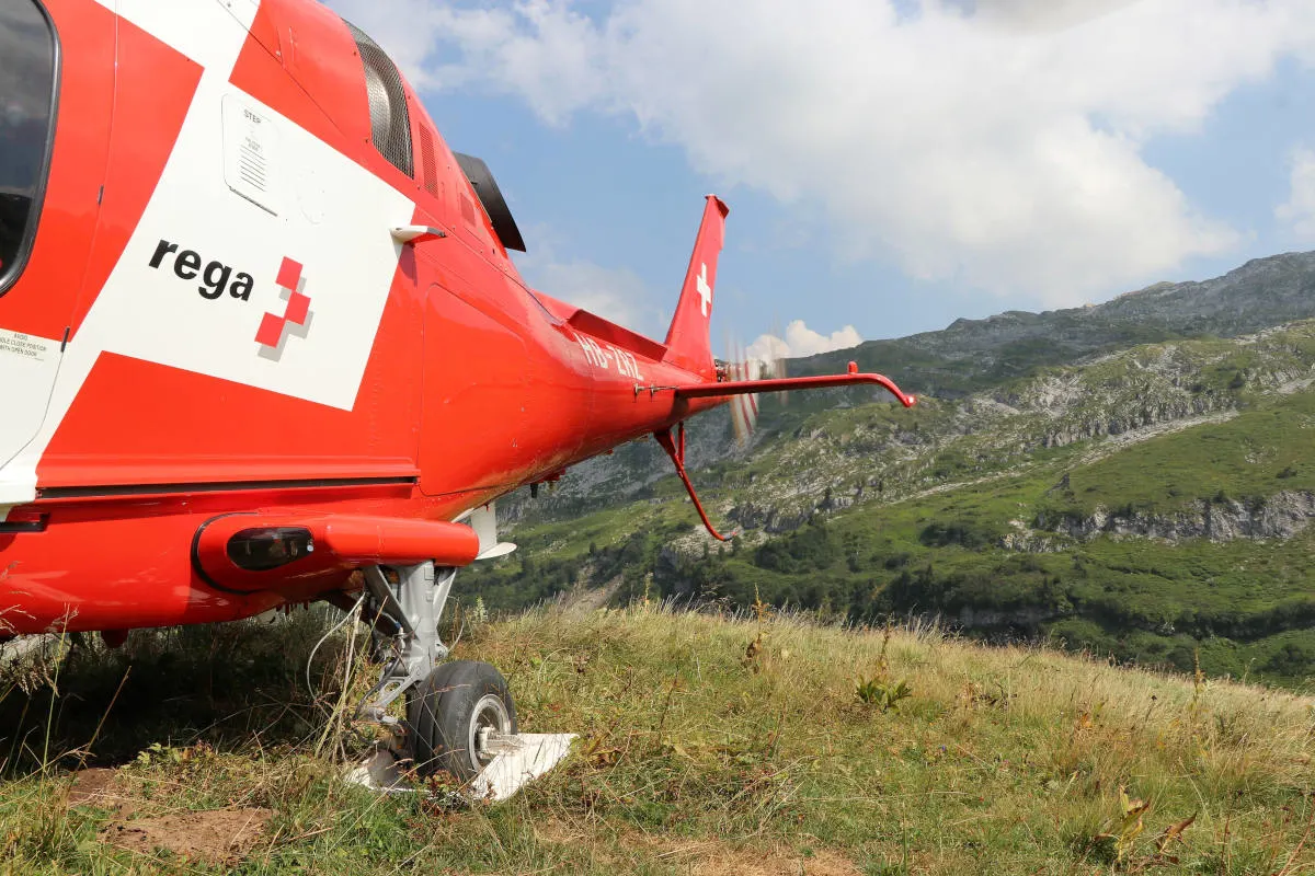 Ein roter Rega-Helikopter ist auf einer grasbedeckten Wiese in den Alpen gelandet. Der Himmel ist klar mit einigen Wolken im Hintergrund.
