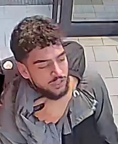 Ein junger Mann mit Bart und lockigem Haar wird von einer Überwachungskamera aufgenommen. Er trägt ein offenes Hemd in einem Gebäude.