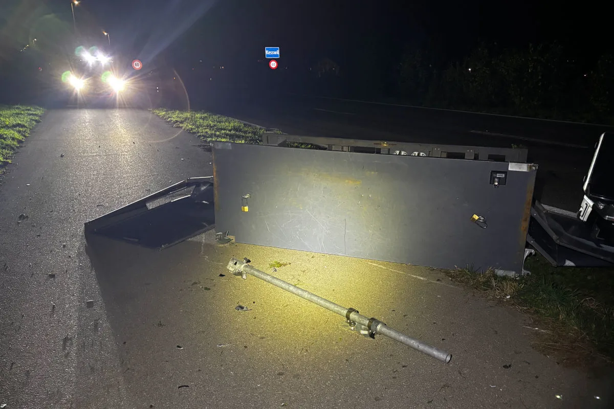 Ein umgestürzter Gegenstand blockiert einen Gehweg bei Nacht. Ein Auto beleuchtet die Szene mit seinen Scheinwerfern.