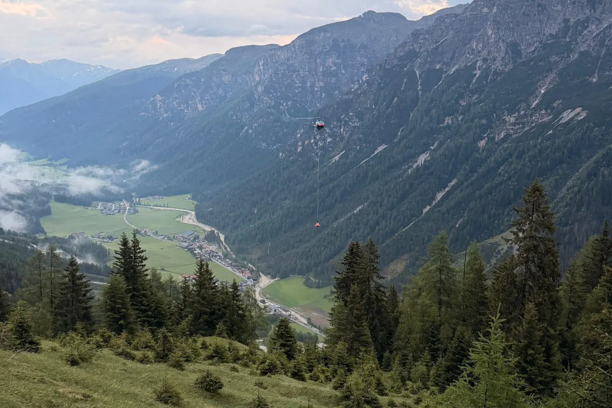 Ein Hubschrauber fliegt in der Mitte des Bildes hoch über einem bewaldeten Gebirgstal und transportiert eine Last darunter. Die Umgebung zeigt ein weitläufiges Tal mit kleinen Siedlungen.