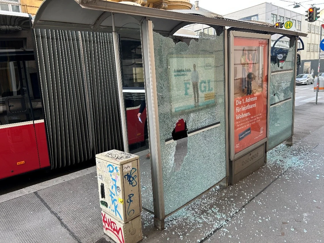 Eine Bushaltestelle mit stark beschädigter Glasscheibe und einem roten Bus im Hintergrund. Glassplitter liegen auf dem Boden.