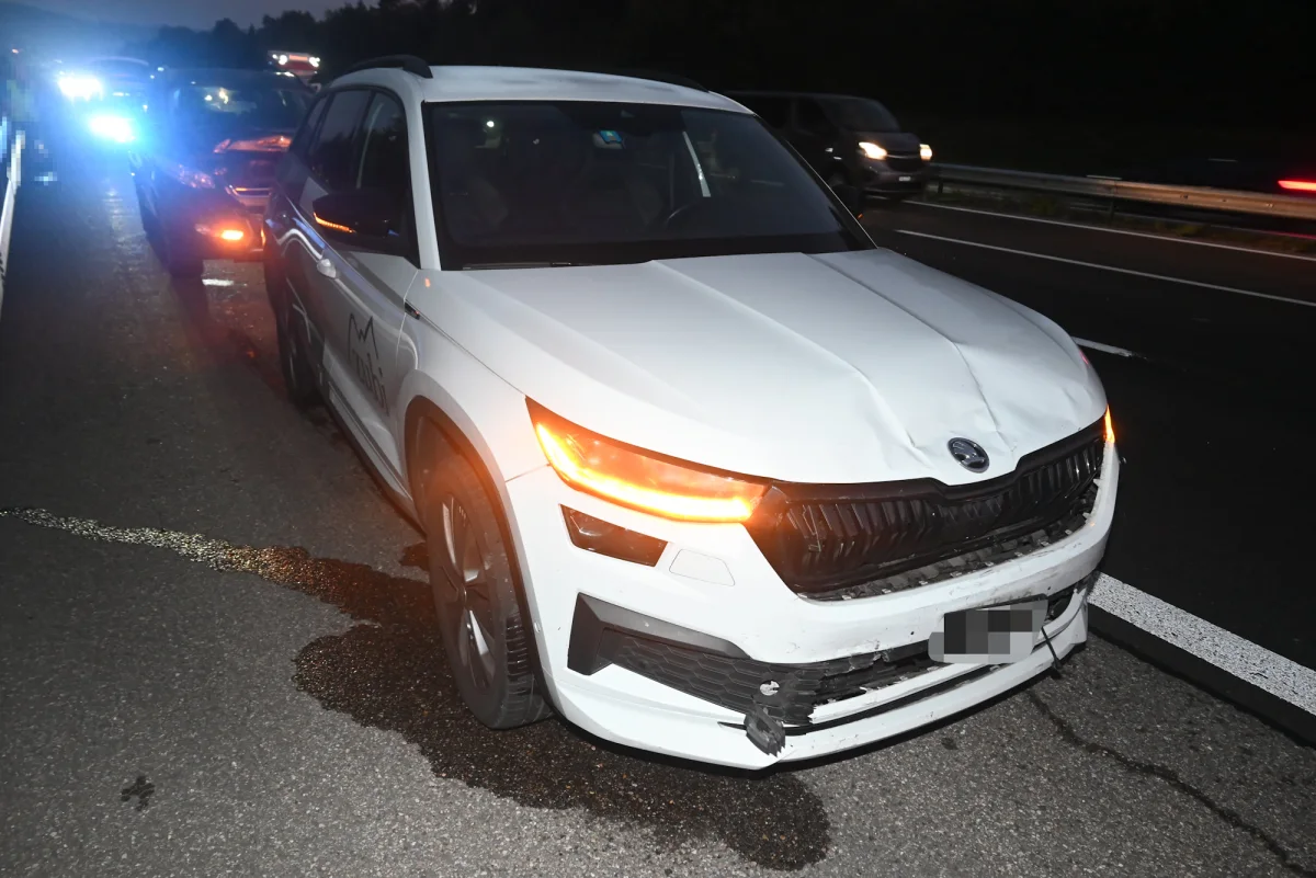 Ein weißes Auto mit sichtbaren Schäden an der Front steht auf einer Autobahn. Im Hintergrund sind weitere Fahrzeuge zu sehen.