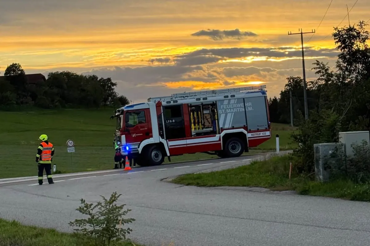 Ein rotes Feuerwehrfahrzeug mit der Aufschrift 'Feuerwehr St. Martin' steht auf einer Landstraße, während ein Beamter mit Warnweste danebensteht. Der Himmel ist mit einem Sonnenuntergang im Hintergrund beleuchtet.