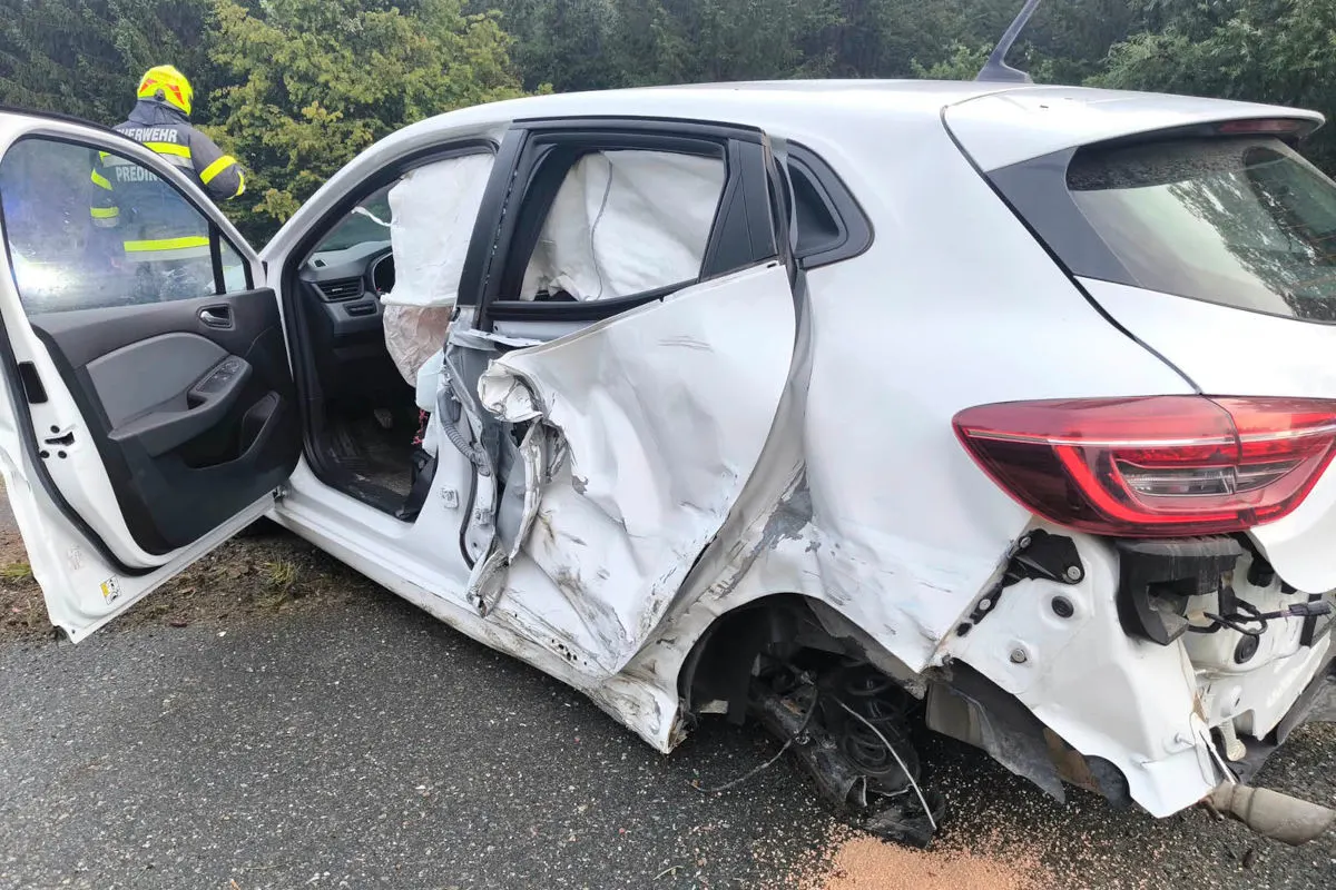 Ein weißes Auto zeigt schwere Seitenschäden und geöffneten Airbags nach einem Unfall. Ein Feuerwehrmann ist im Hintergrund zu sehen.