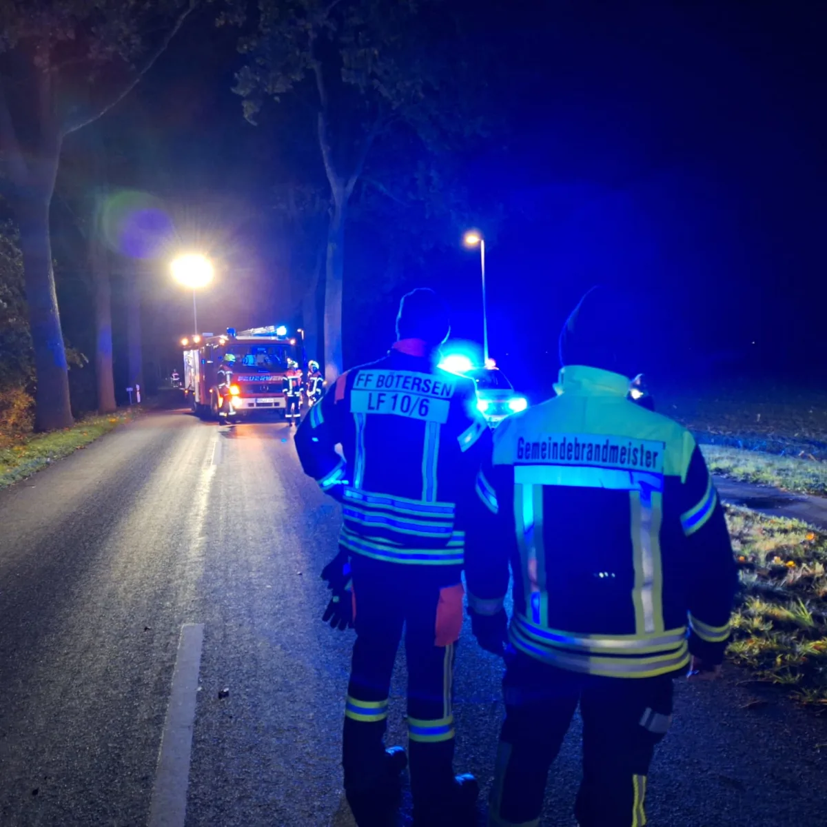 Zwei Feuerwehrleute stehen auf einer Landstraße, beleuchtet von Blaulicht und dem Scheinwerfer eines Feuerwehrfahrzeugs im Hintergrund.