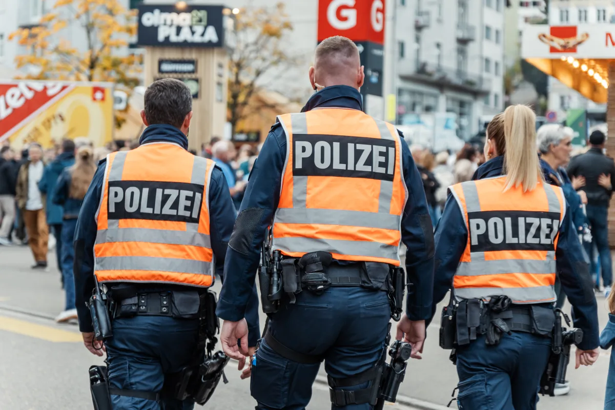Drei Polizisten in orangefarbenen Westen stehen in einer Menschenmenge in der Stadt. Im Hintergrund sind Schilder und Gebäude sichtbar.