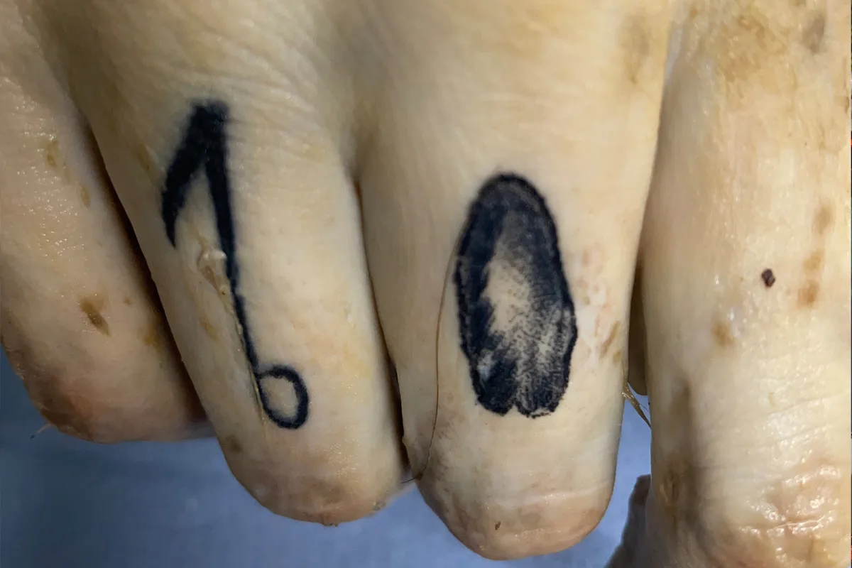 Die Nahaufnahme zeigt zwei Finger, von denen einer eine eins an der Seite tätowiert hat und der andere ein rundes Motiv.