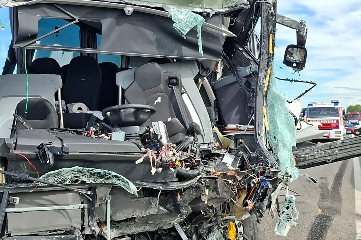 Das Bild zeigt das stark beschädigte Fahrerhaus eines Fahrzeugs mit zerbrochenen Scheiben und freiliegenden Kabeln. Im Hintergrund sind Feuerwehrfahrzeuge zu sehen.
