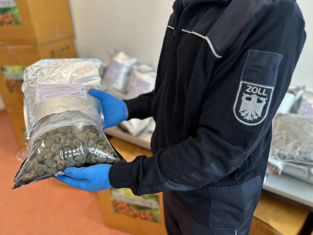 Ein Zollbeamter, erkennbar an der Uniform, hält eine versiegelte Tüte mit einer großen Menge an Substanzen. Im Hintergrund sind weitere Tüten auf einem Tisch zu sehen.