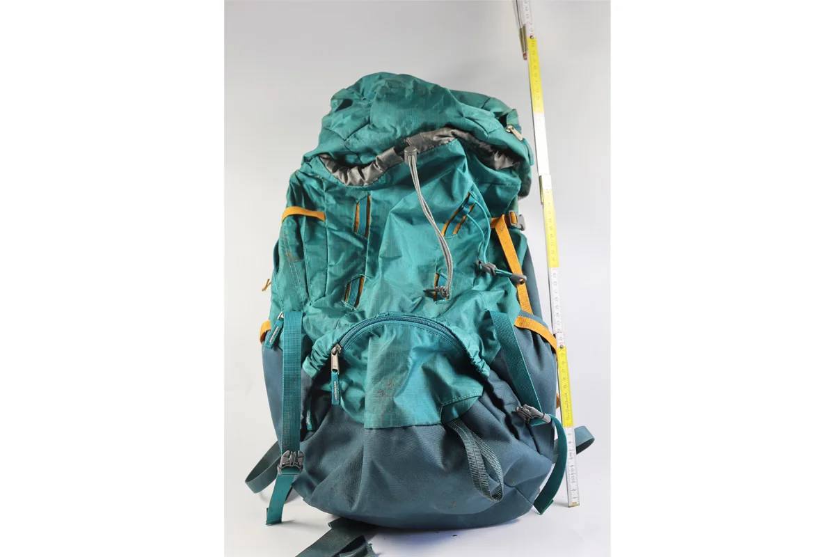 Ein großformatiger, türkisfarbener Wanderrucksack mit gelben Akzenten steht vor einem neutralen Hintergrund. Der Rucksack wird von einem Maßstab flankiert, der seine Größe hervorhebt.