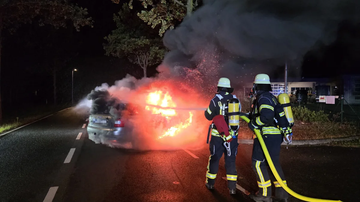 Zwei Feuerwehrleute mit Atemschutzmasken und Schläuchen löschen bei Nacht ein Auto, das auf einer Straße Feuer gefangen hat.