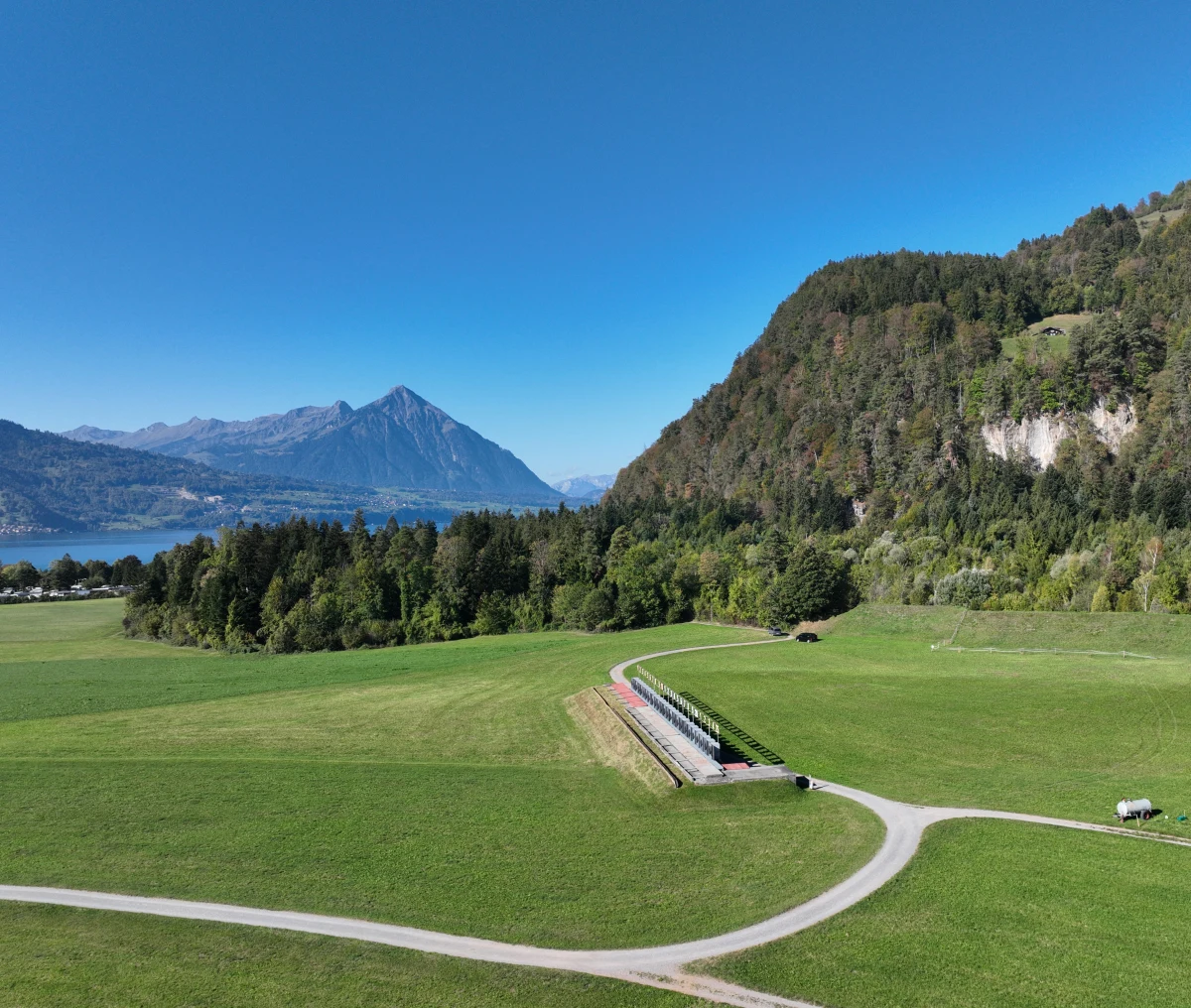 Eine weite grüne Wiese erstreckt sich vor einer Hügelkette. Im Hintergrund sieht man Berge und einen See unter blauem Himmel.