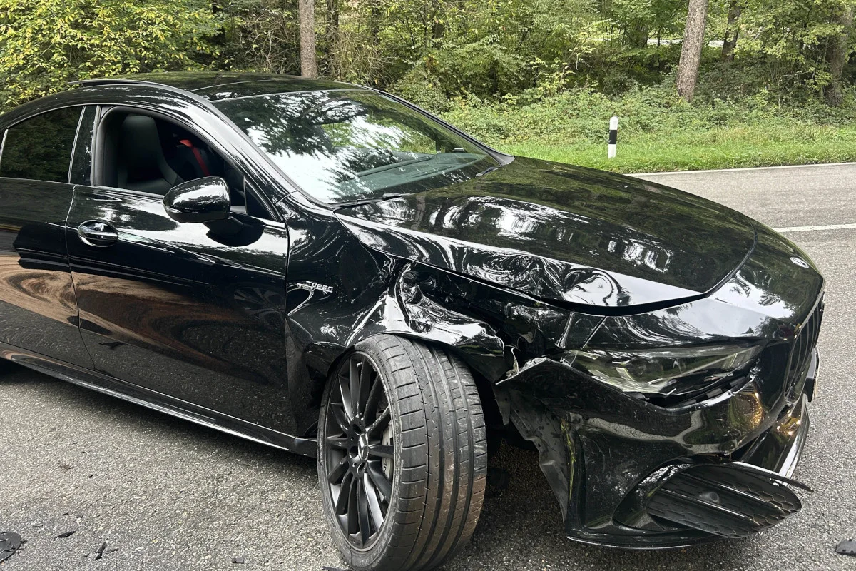 Ein schwarzes Auto mit stark beschädigter Front und einem verdrehten Vorderrad steht auf einer asphaltierten Landstraße. Im Hintergrund ist Wald zu sehen.